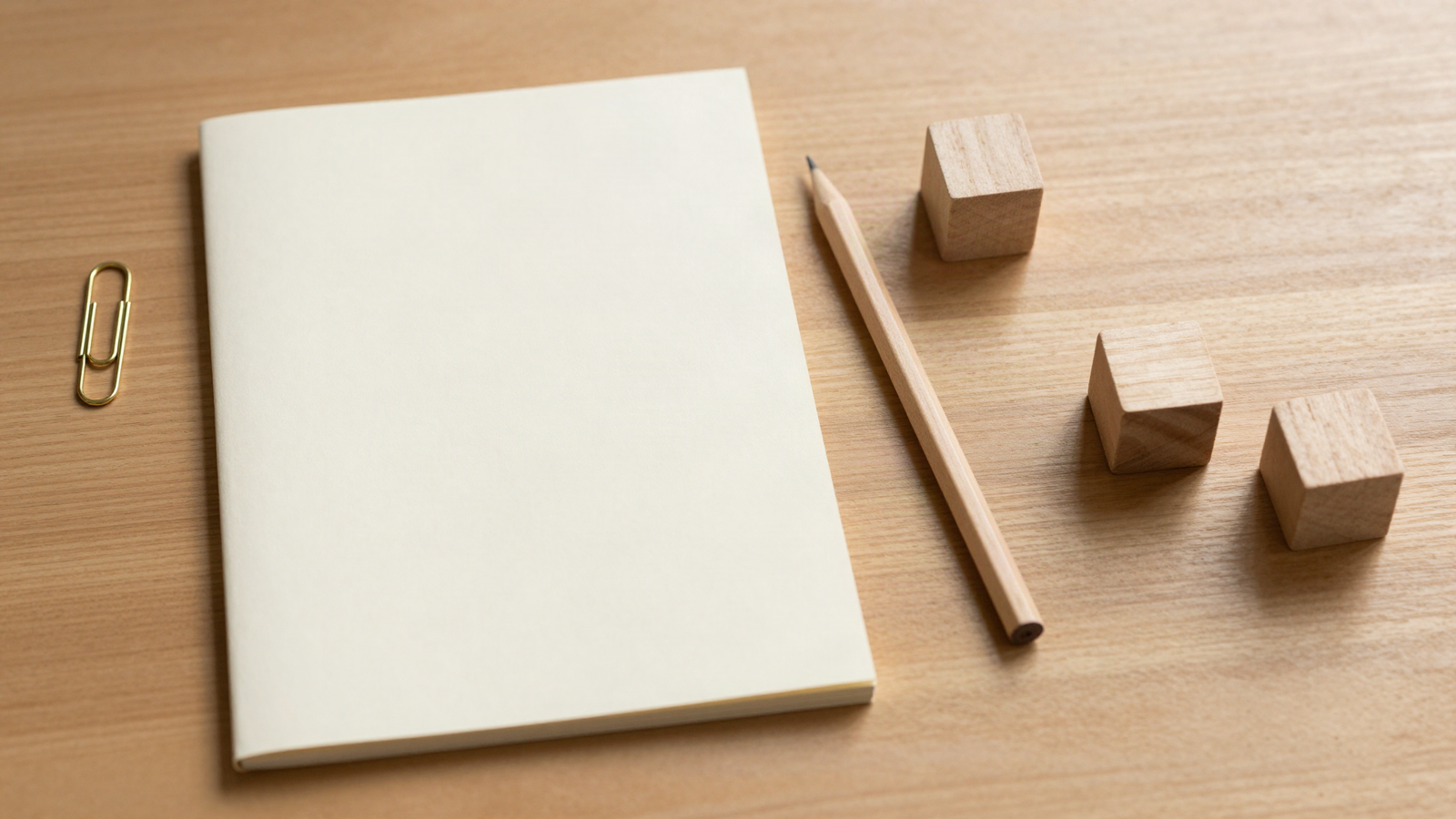 Blank cream notepad, sharpened pencil, brass paperclip and three small wooden blocks arranged on a warm oak desk